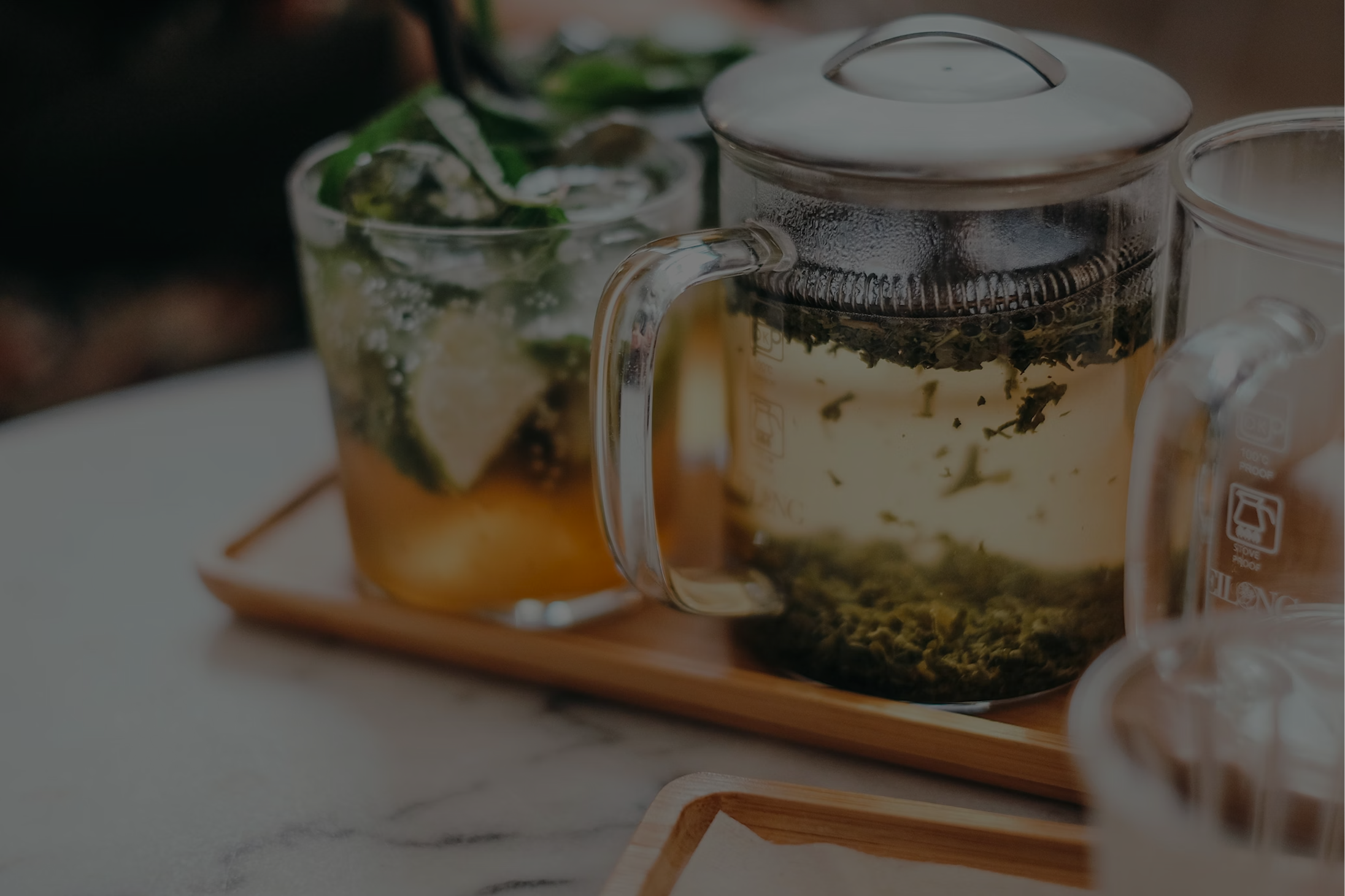 Glass teapot with steeping green tea leaves and a glass of iced tea with lime and mint on a wooden tray.