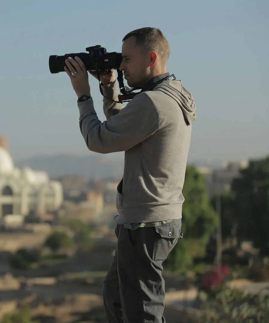 Man wearing a gray hoodie and dark pants holding a camera and taking a photo outdoors with a blurred cityscape in the background.