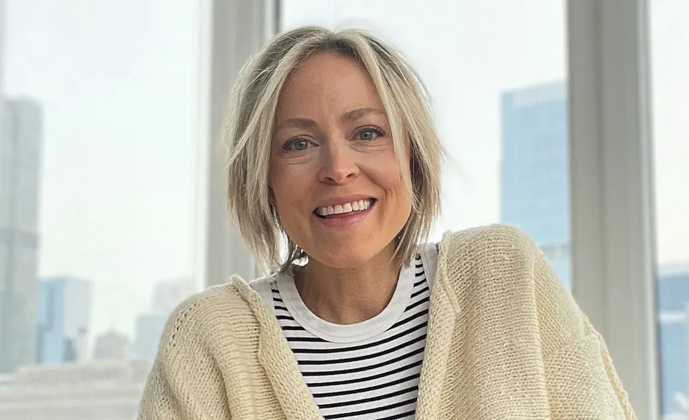 Smiling woman with short blonde hair wearing a striped shirt and cream knitted sweater, with city buildings visible through large windows in the background.