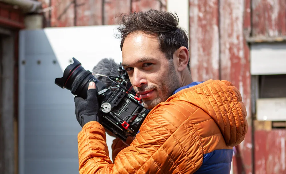 Man in an orange jacket holding a professional video camera outdoors near a red wooden building.