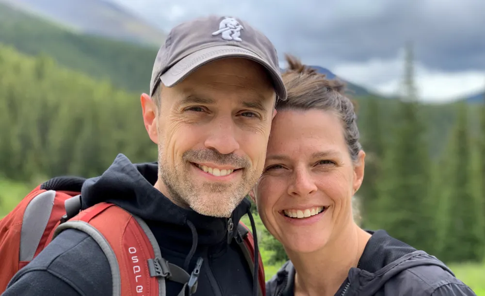Smiling man and woman outdoors with forest and mountains in the background, man wearing a cap and a red backpack.