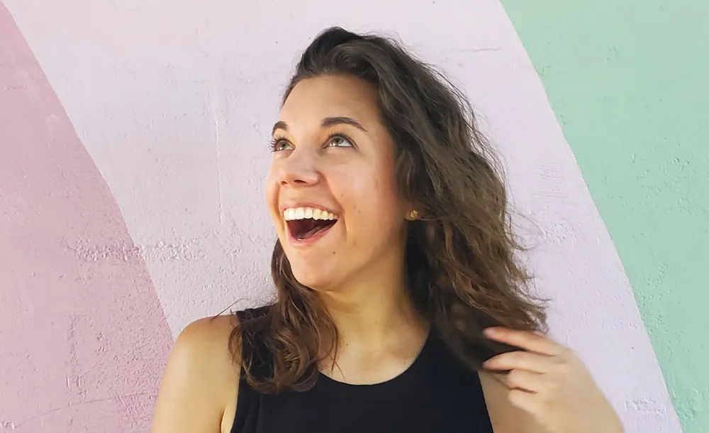 Young woman with curly brown hair smiling and looking upwards against a pastel-colored wall.