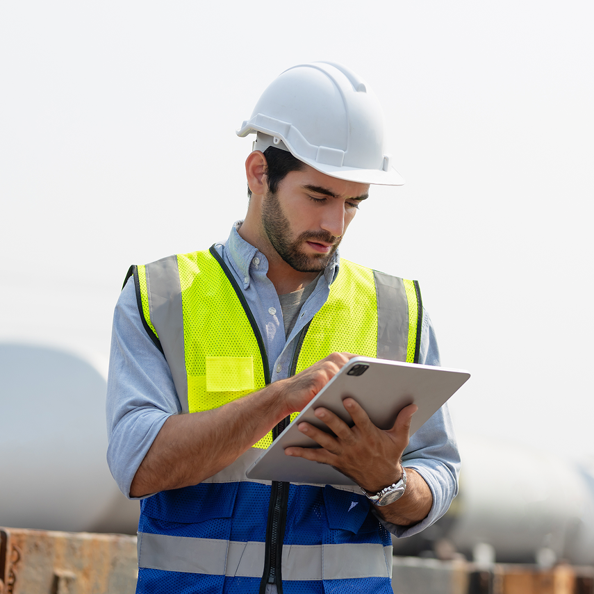 Construction worker wearing a yellow hard hat and high-visibility shirt using a tablet outdoors near a building site.