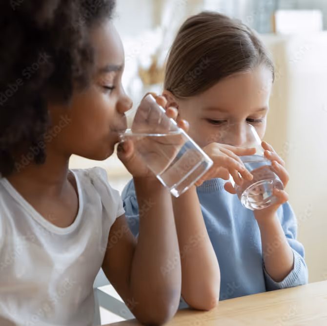 Two young girls sitting at a table drinking water from clear glasses.