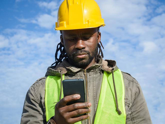 Construction worker wearing a yellow hard hat and green safety vest looking at a smartphone against a blue sky.