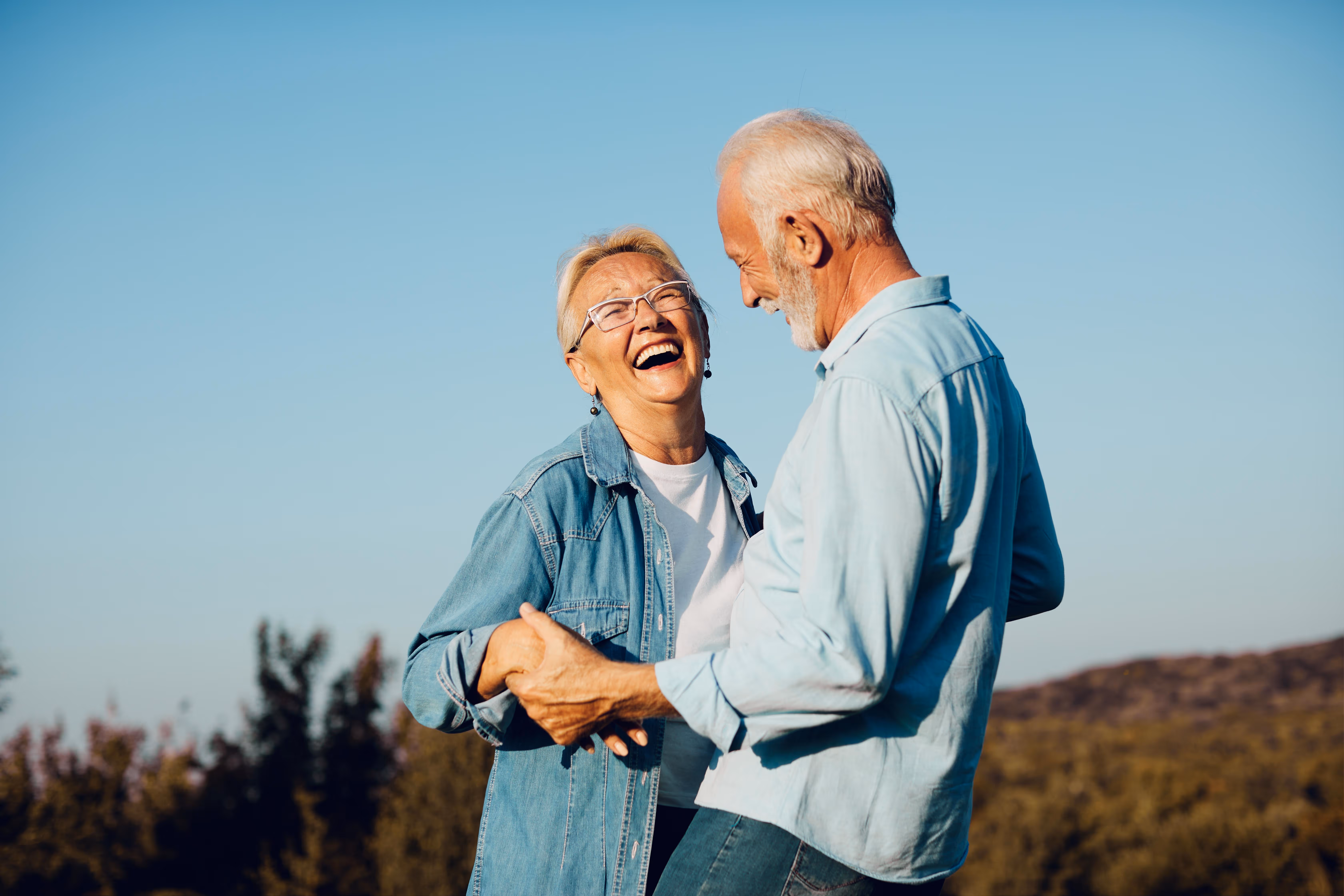 Elderly couple holding hands and laughing outdoors under clear sky.