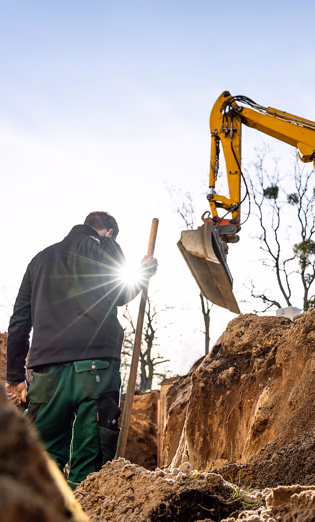 Construction worker holding a shovel standing next to a large excavator digging a trench in the ground.