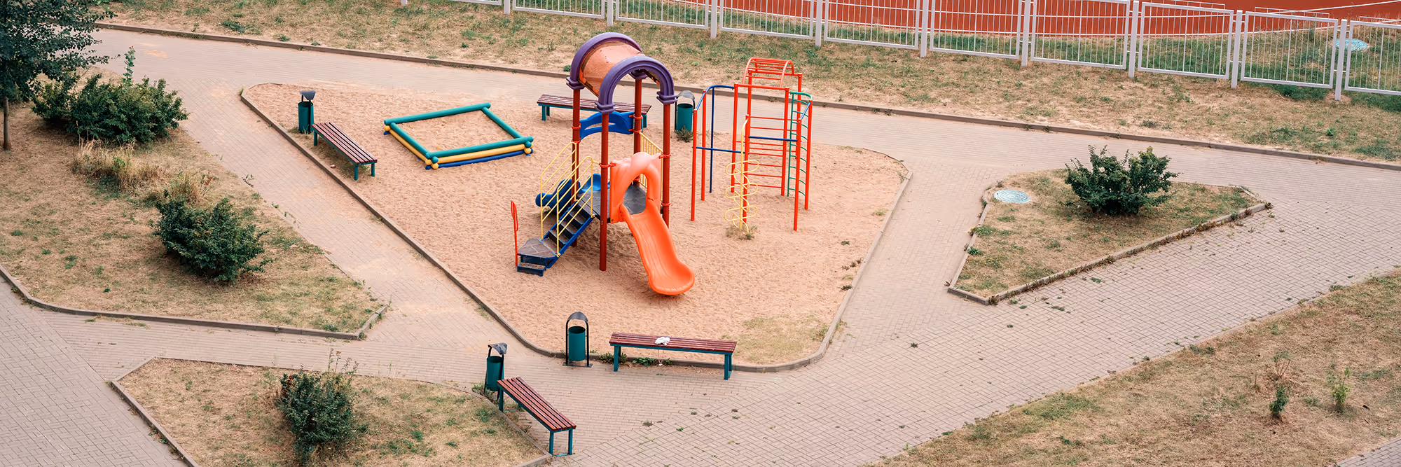 Outdoor playground with an orange slide, climbing structures, benches, and a sandpit surrounded by paved walkways and patches of grass with shrubs.