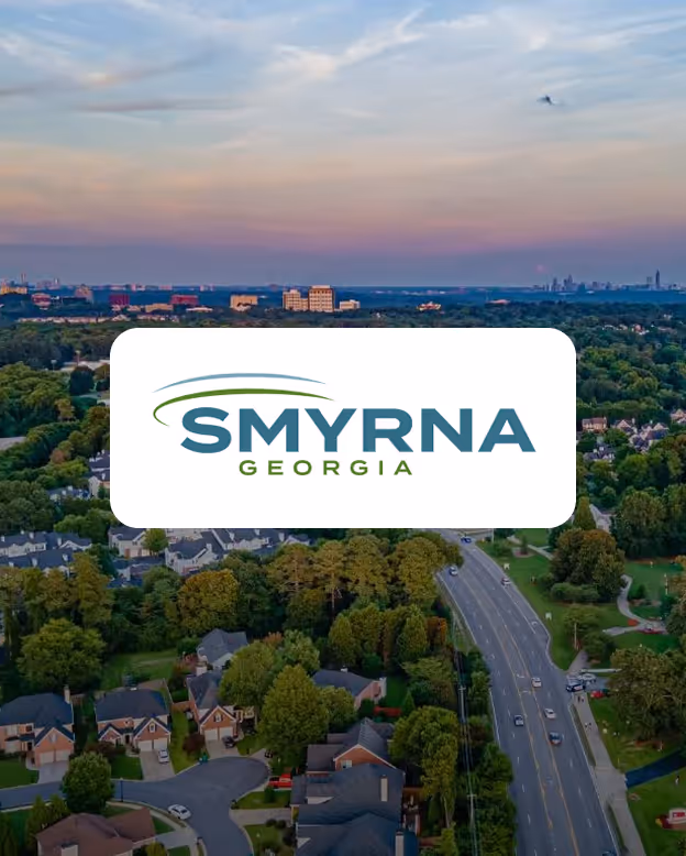 Aerial view of Smyrna, Georgia residential area with houses, trees, and a main road leading towards city buildings under a cloudy sky at sunset, with Smyrna Georgia logo overlayed.