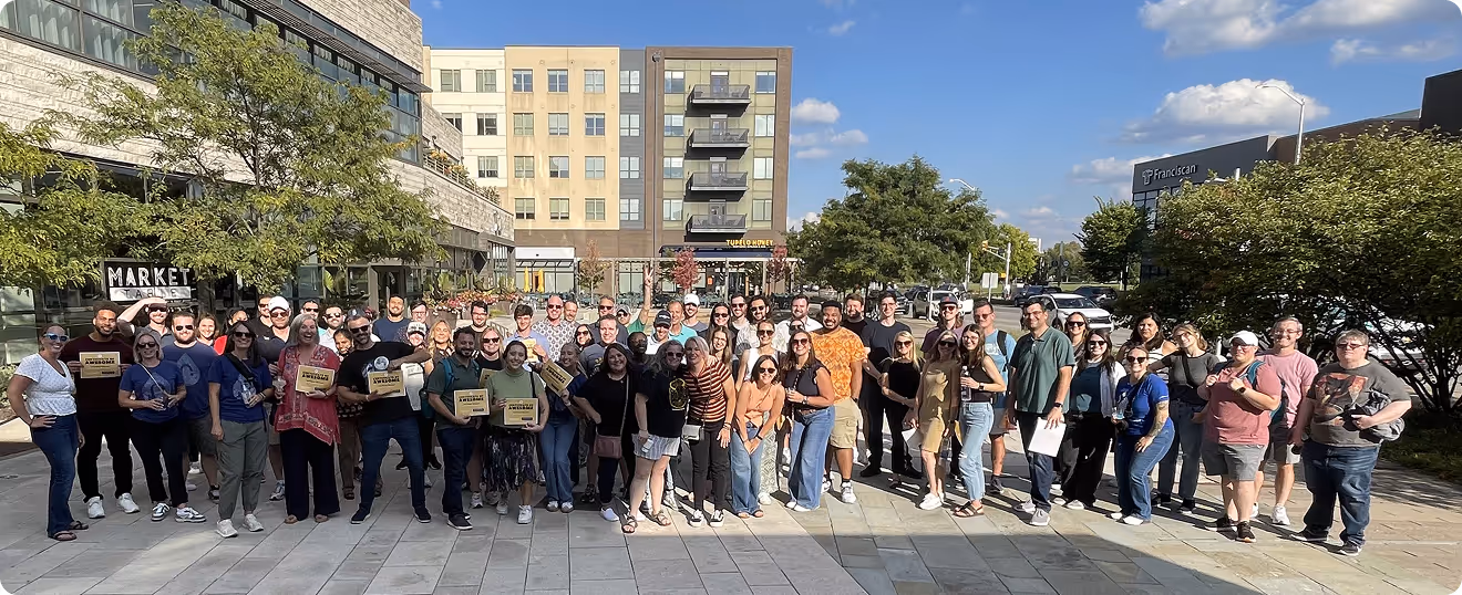 Large group of people posing outdoors on a sunny day in an urban plaza with buildings and trees in the background.