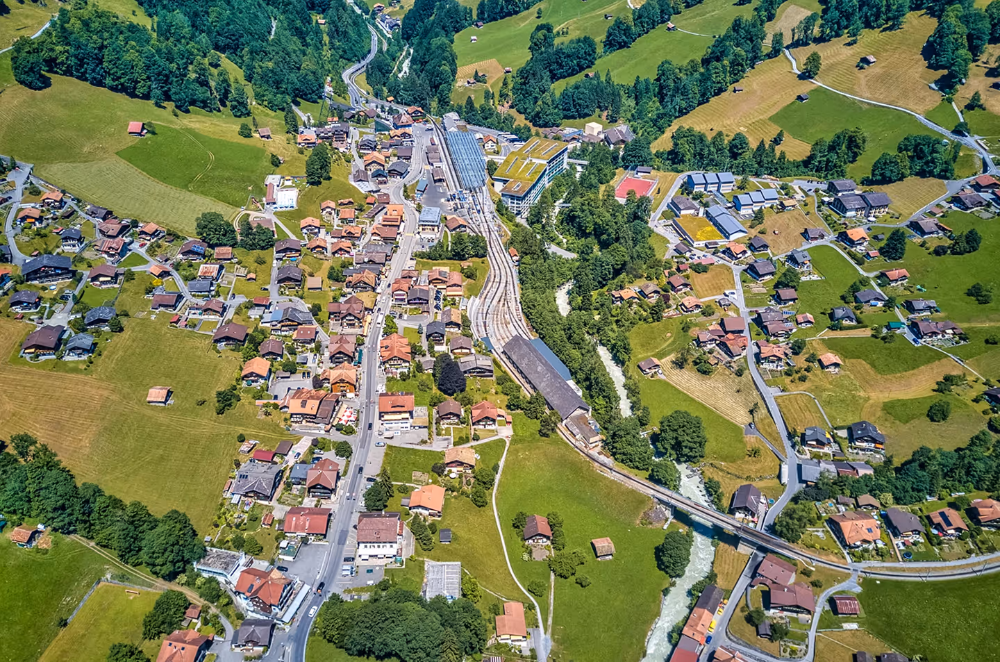 Aerial view of a village with clustered houses, roads, green fields, trees, a river, and a railway station with train tracks.