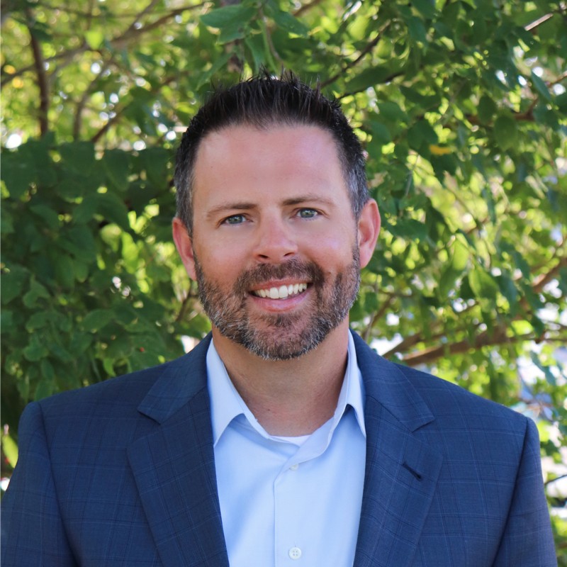 Smiling man with short dark hair, beard, and blue eyes wearing a blue suit jacket and light blue shirt standing outdoors with green leafy background.