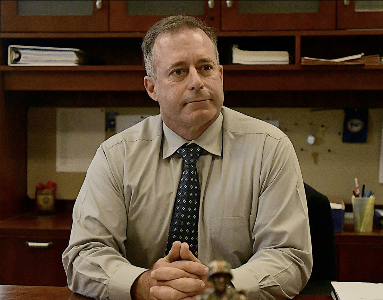 Man in a beige shirt and dark patterned tie sitting at a desk with hands clasped, with shelves and office supplies in the background.