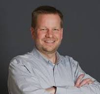 Smiling man with short hair wearing a light blue button-up shirt against a dark gray background.