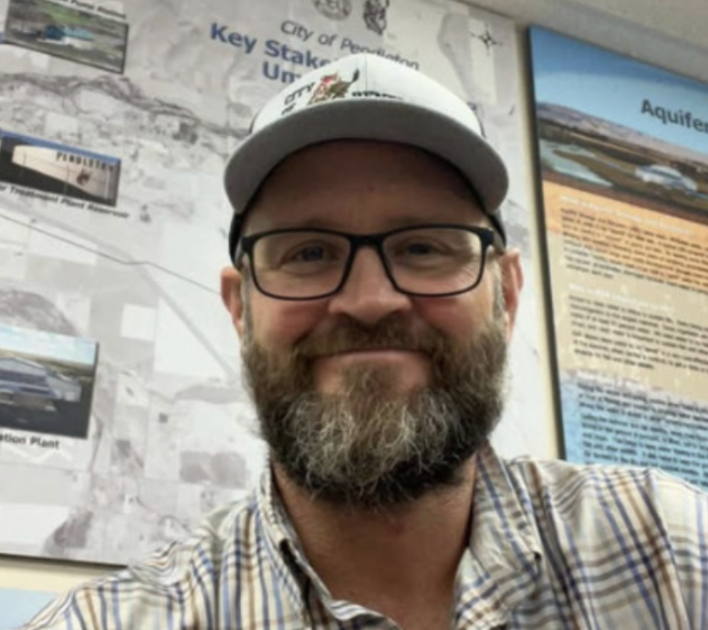 Bearded man wearing glasses and a white baseball cap smiling in front of informational posters on a wall.