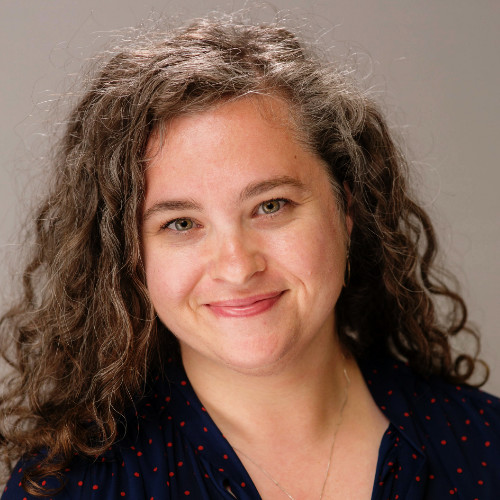 Smiling woman with curly brown hair wearing a navy blouse with red polka dots against a plain background.