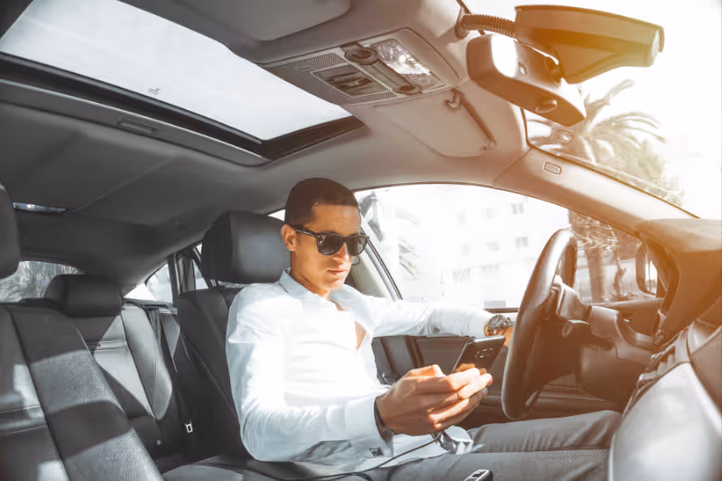 A man in white collared shirt and sunglasses is texting while driving his car