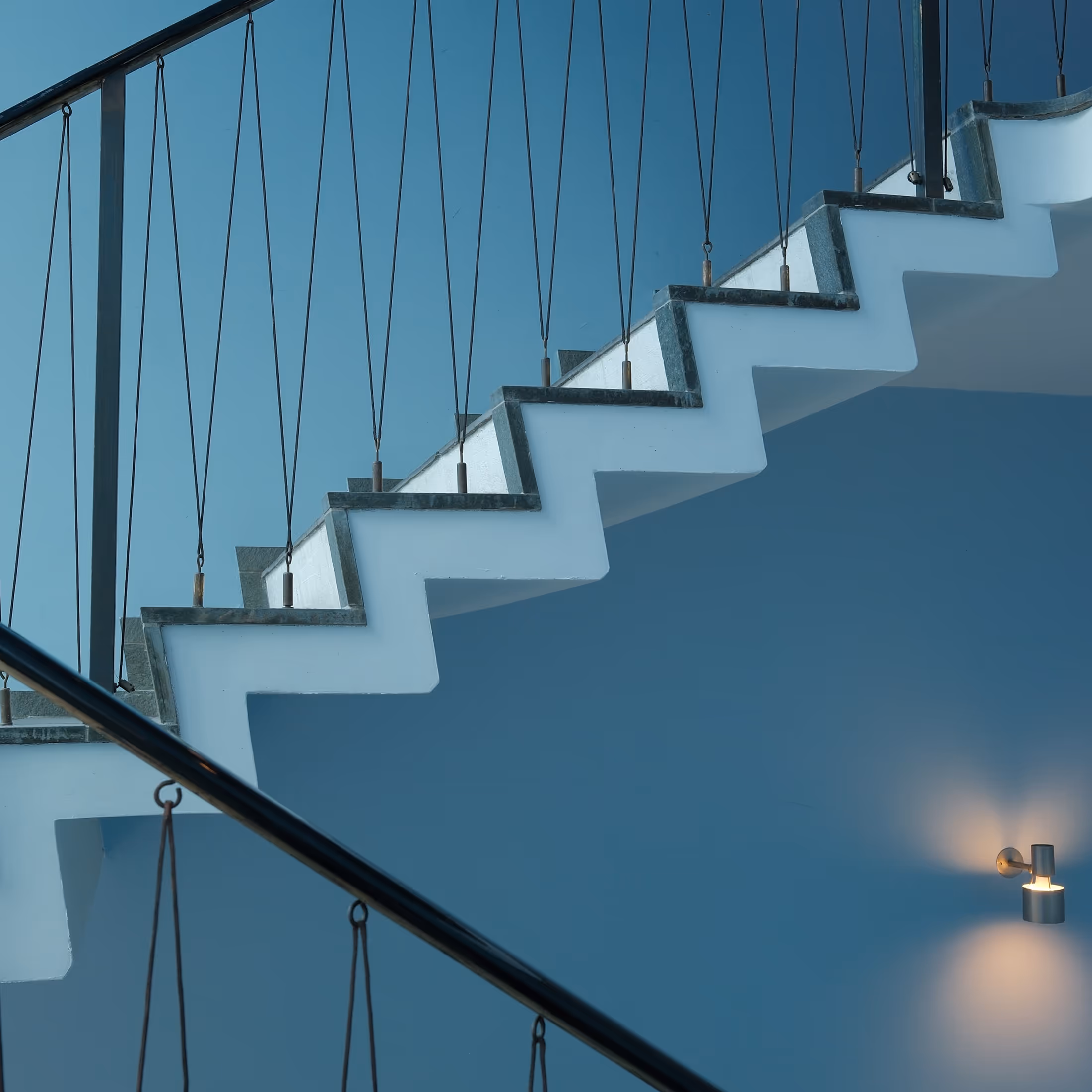 Modern white staircase with dark metal railing and vertical cables against a blue wall with a glowing wall light.