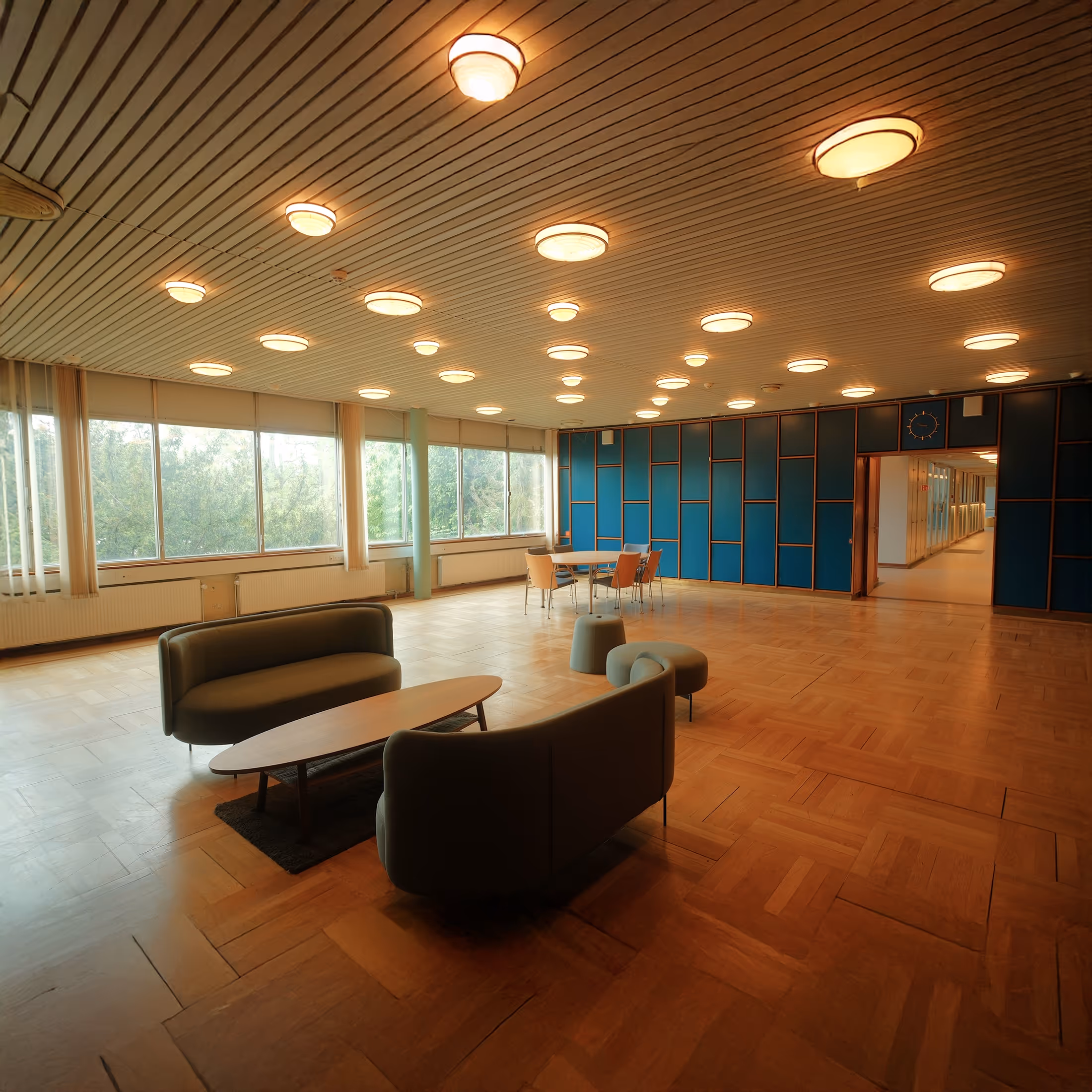 Spacious lounge area with wooden floor, large windows, two curved sofas around a coffee table, and a dining table with chairs by a blue paneled wall.
