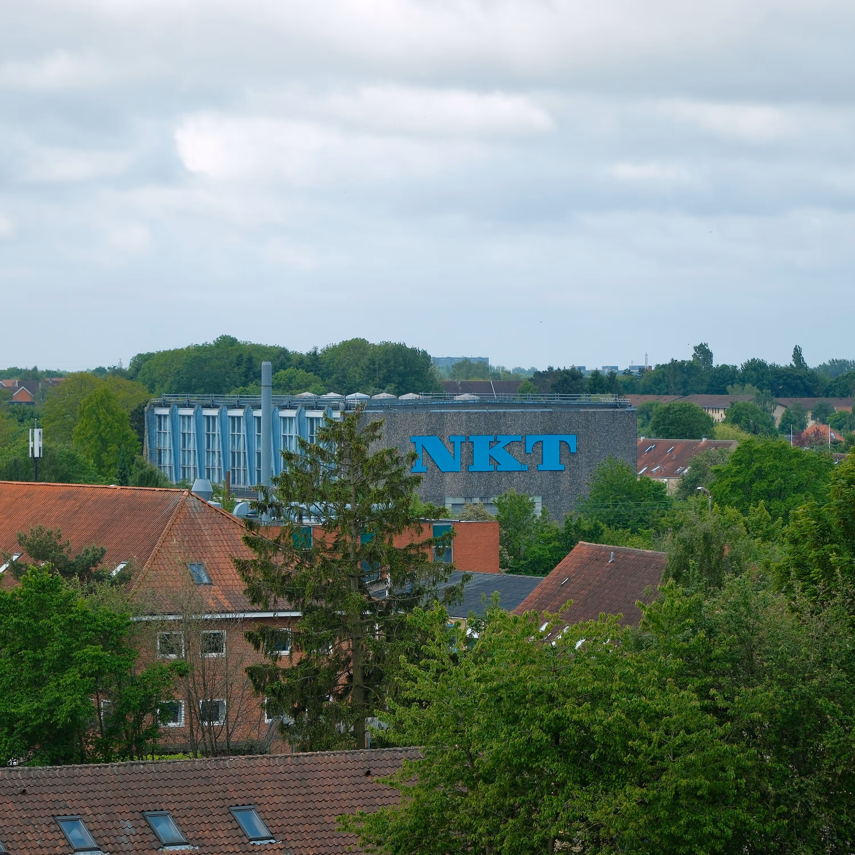 View of a building with blue letters NKT on the facade surrounded by green trees and red-roofed houses under a cloudy sky.