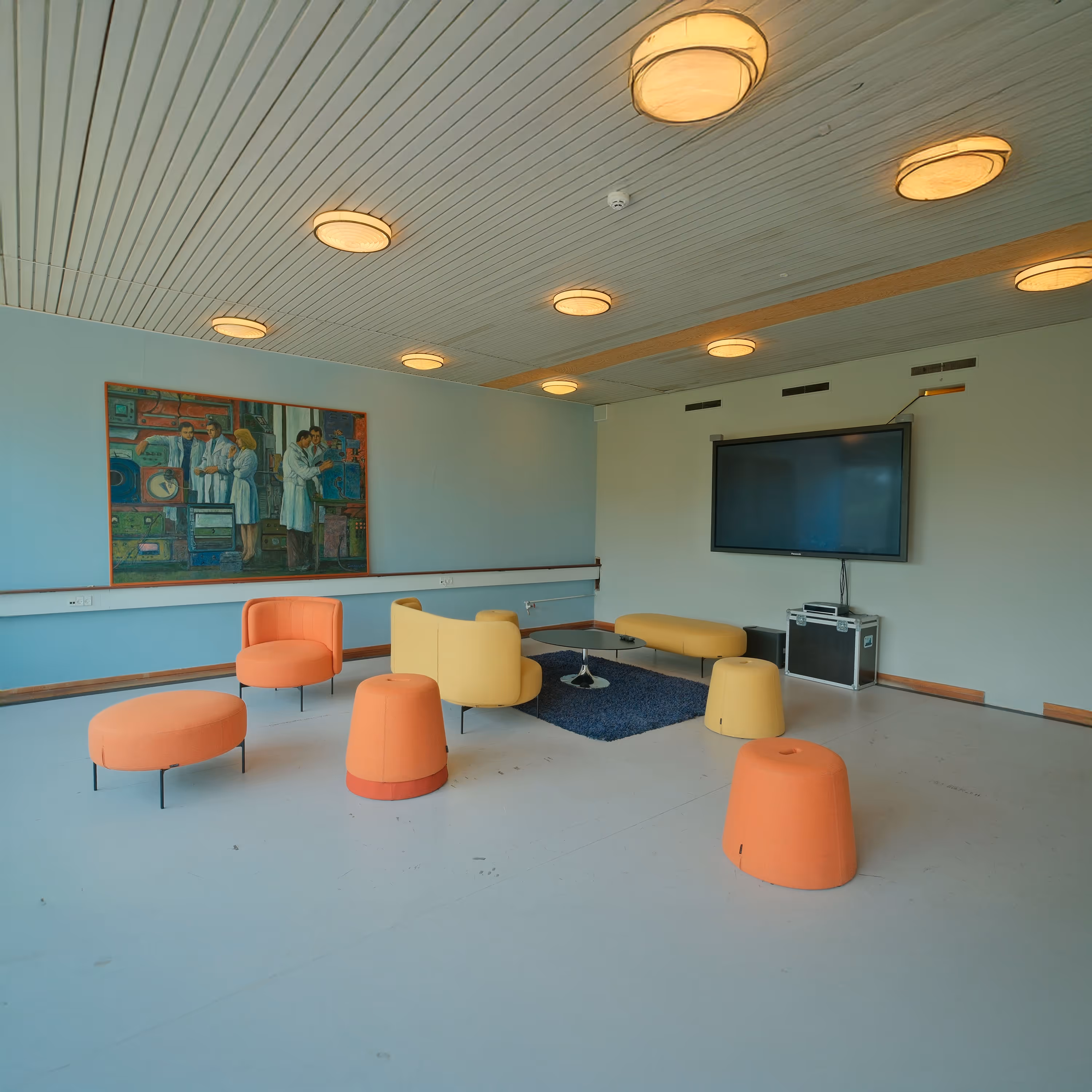 Modern lounge room with orange and yellow seating, a blue rug, wall-mounted TV, and a painting of scientists on the wall.