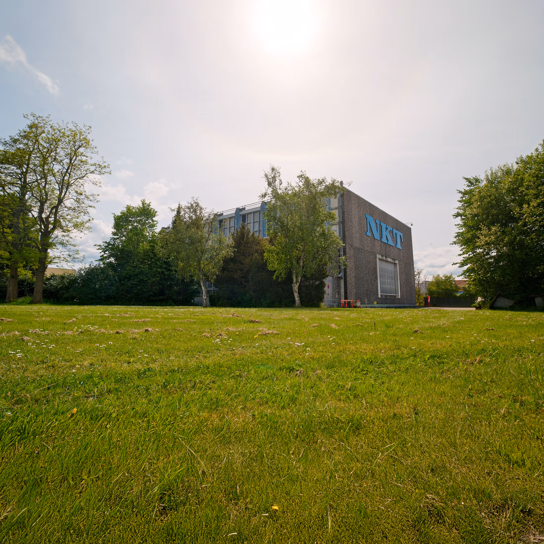 Building with NKT logo surrounded by green trees and grass under a sunny sky.