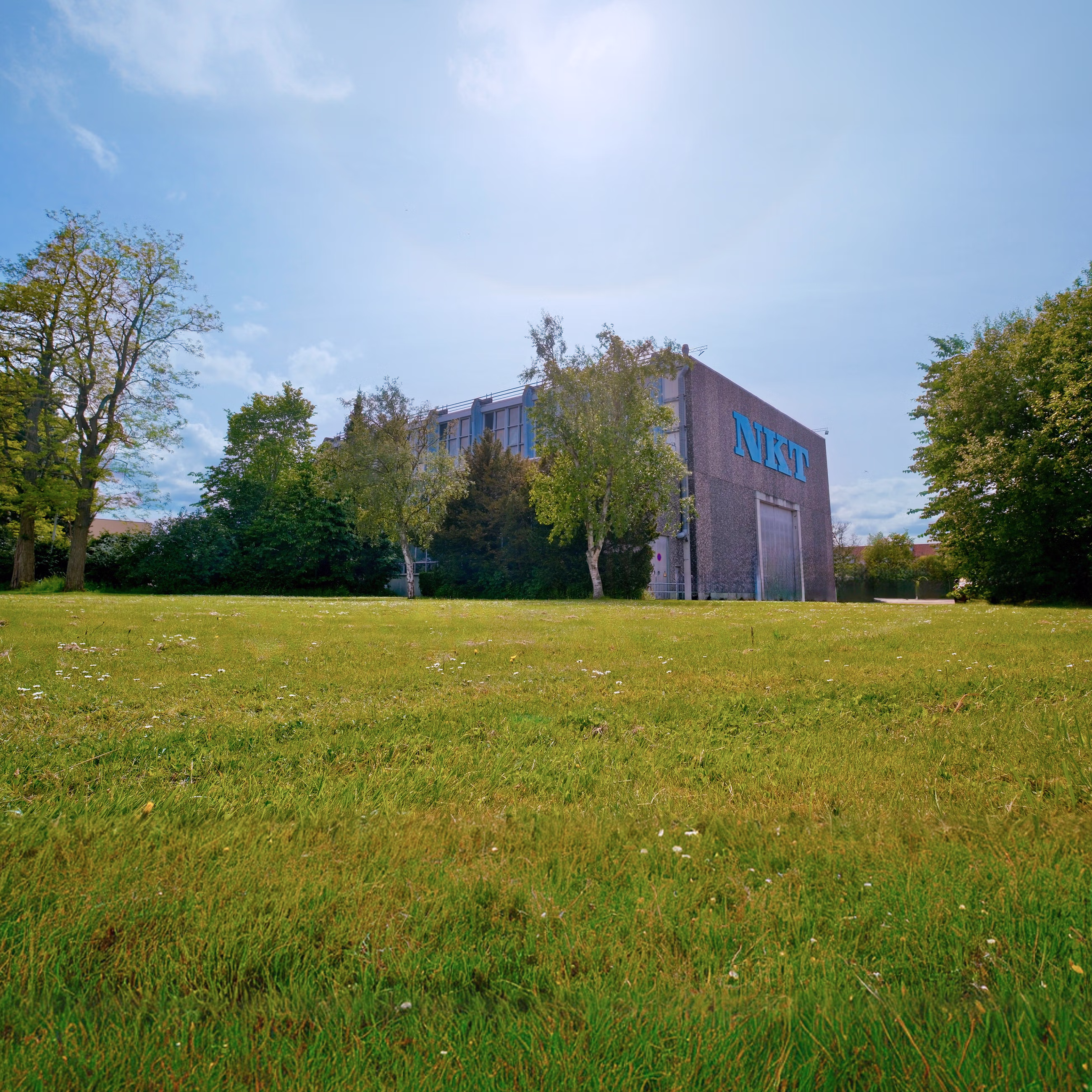 Building with NKT logo surrounded by green trees and grass under a sunny sky.