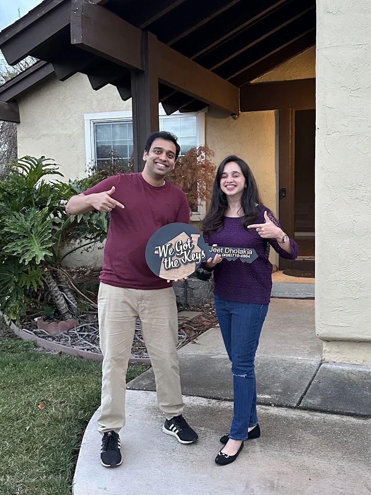 Smiling man and woman holding a sign that says We Get the Keys in front of a house entrance.