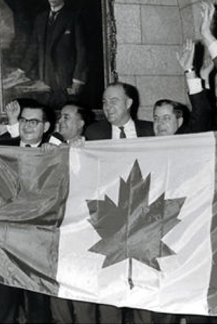 Black and white photo of four men in suits holding a Canadian flag with a maple leaf, raising their hands in celebration.