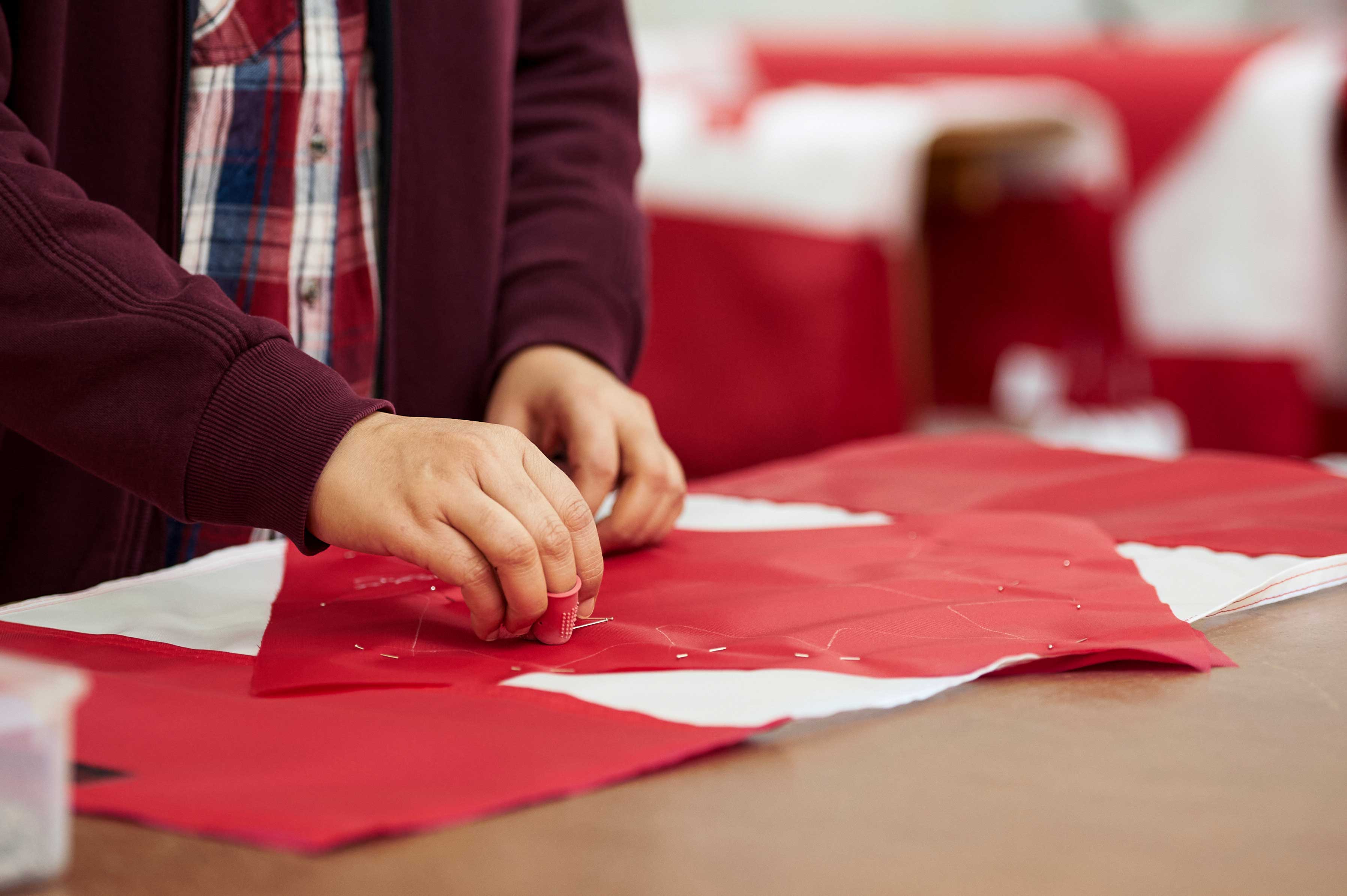 Person hand-pinning red and white fabric pieces on a table, wearing a plaid shirt and maroon sweater.