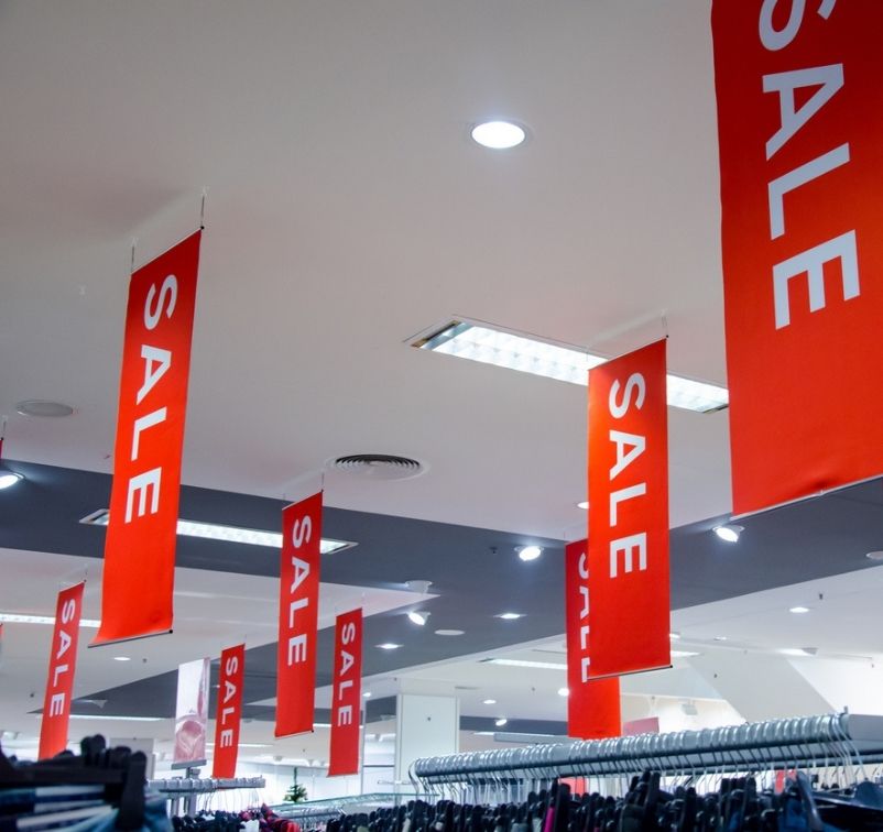 Multiple red sale banners hanging from the ceiling above clothing racks in a retail store.