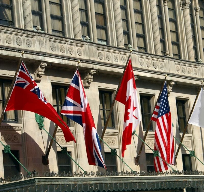 Flags of the United Kingdom, Canada, and the United States displayed on poles outside a stone building with large windows.
