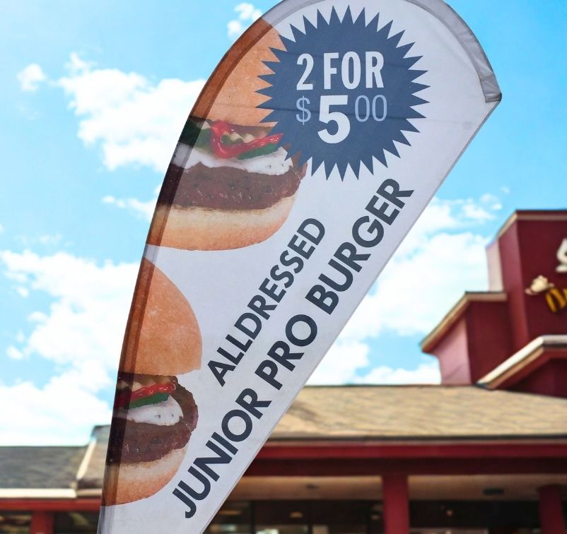 Promotional flag for two all-dressed junior pro burgers priced at $5.00 each, with a fast food restaurant building in the background.