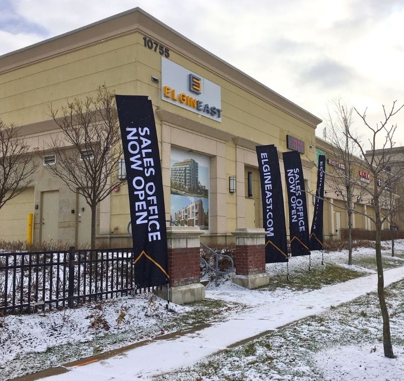 Exterior of Elgin East building with flags announcing sales office now open on a snowy sidewalk.