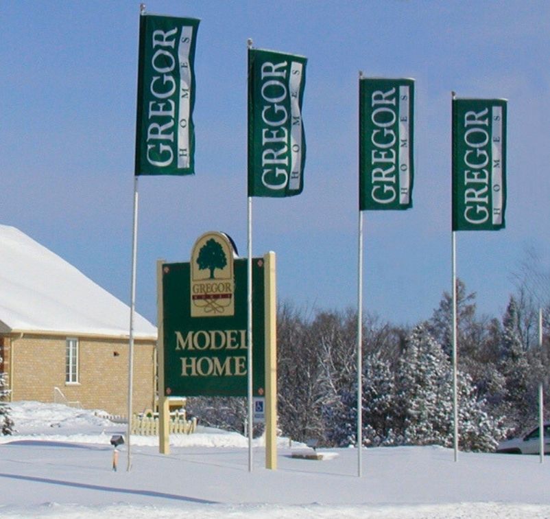 Snow-covered yard with four green flags and a sign reading 'Gregor Homes Model Home' in front of a brick house.