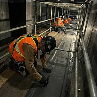 Ouvriers spécialisés d'Elastomont effectuant l'installation de revêtement 'rubber lining' sur un chantier industriel au Québec (Montréal/Fermont). Vue de trois travailleurs en équipement de protection individuelle (EPI) appliquant des feuilles de caoutchouc au sol dans un espace confiné sous des échafaudages.