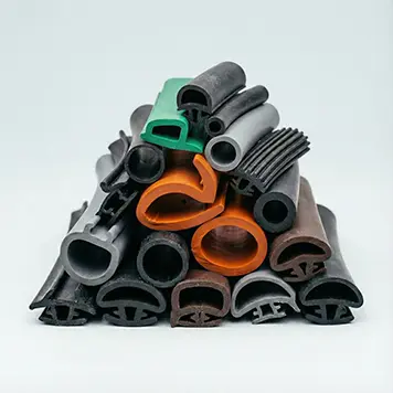 Une pile de joints en caoutchouc extrudés de formes et de tailles variées, incluant des pièces noires, grises, orange et une verte, sur un fond gris pâle. Ces produits sont typiques des extrusions fabriquées par des entreprises comme Elastomont à Montréal, Québec.
