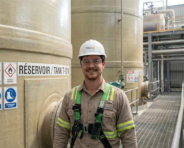 Photo d'un responsable de l'approvisionnement d'une firme d'ingénierie basée à Montréal, Québec, posant devant un grand réservoir industriel identifié comme ESPACE CLOS et DANGER. Il témoigne de l'efficacité des solutions de revêtement en caoutchouc d'Elastomont pour la protection de leurs équipements critiques.