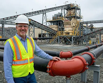 Un directeur de maintenance souriant, portant un casque et un gilet de sécurité haute visibilité, pose devant une nouvelle ligne de tuyauterie de 8 pouces dans une usine de traitement de mine de diamants. Il touche un coude rouge vif équipé du système Victaulic XL rubber lining, utilisé conjointement avec la technologie Elastomont pipe lining pour remplacer les anciens tuyaux en HDPE. L'infrastructure industrielle de la mine est visible en arrière-plan.