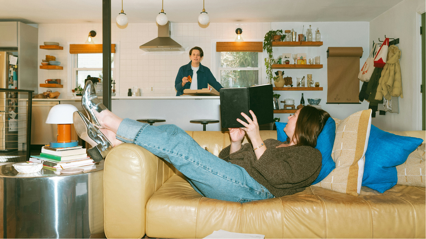A woman lying on a sofa reading a notebook, with a man standing in the kitchen in the background of a cozy home