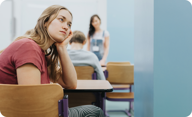 Adolescente en aula escolar sentada con expresión pensativa mientras escucha una profesora al fondo