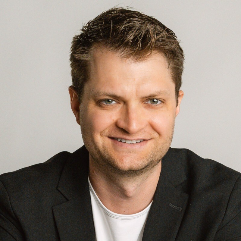 Smiling man with short brown hair wearing a black blazer and white shirt against a plain background.