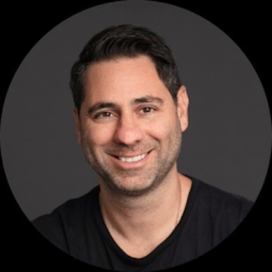 Smiling man with short dark hair wearing a black shirt against a dark background.