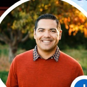 Smiling man wearing a red sweater and checkered shirt standing outdoors with blurred trees in the background.