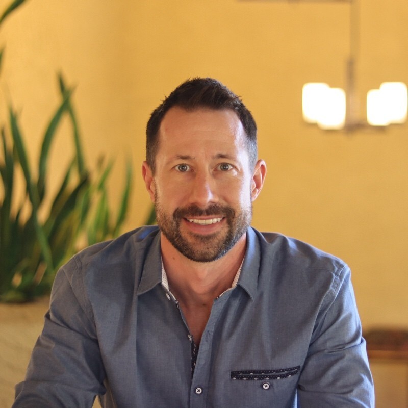 Smiling man with short dark hair and beard wearing a blue button-up shirt sitting indoors with plants and a light fixture in the background.