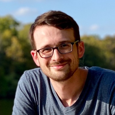 Smiling man with glasses and short brown hair outside with trees and blue sky in the background.