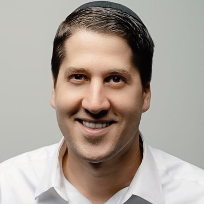 Smiling man with short dark hair wearing a white shirt and a dark kippah against a plain light gray background.