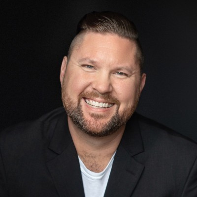 Smiling man with short hair and beard wearing a black blazer and white shirt against a dark background.