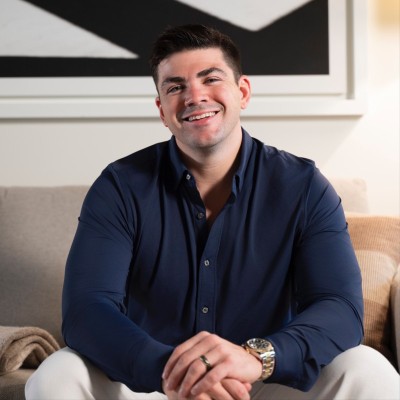 Smiling man with dark hair wearing a navy blue shirt and a watch sitting on a beige couch.