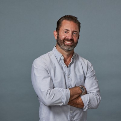 Smiling man with folded arms wearing a light gray button-up shirt against a plain gray background.